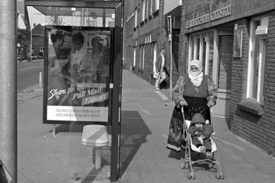 853648 Afbeelding van een migrant met een kind in een wandelwagentje in de Kanaalstraat te Utrecht, met links een abri ...